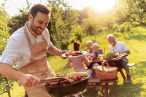 Familie beim grillen. Mann steht am Grill. Zwei Frauen, ein Mann und ein Kind sitzen am Tisch in einem Garten.