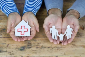 Eine aufgehaltene Kinder- und eine aufgehaltene Erwachsenenhand halten ein Papierhaus und eine Papierfamilie.