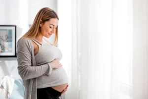 Schöne schwangere Frau berührt ihren Bauch, wenn sie zu Hause am Fenster steht