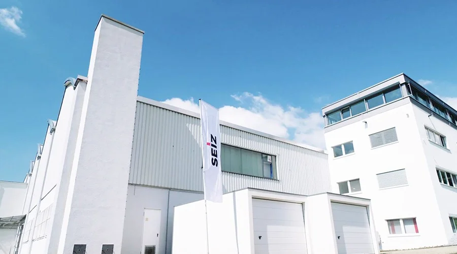Modernes Industriegebäude mit weißer Fassade und SEIZ-Fahne vor blauem Himmel.