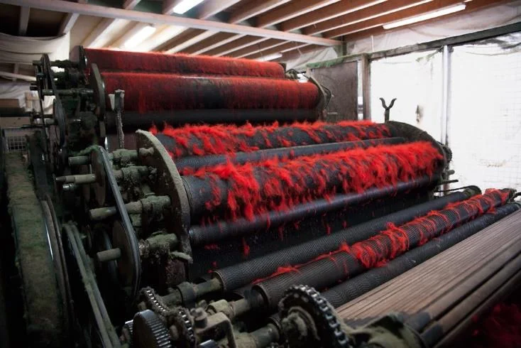 Große industrielle Textilmaschine mit rot gefärbten Fasern auf rotierenden Walzen in einer Produktionshalle.